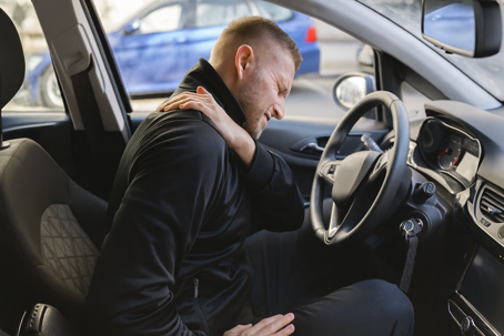 Man sitting in the driver’s seat of a car, wincing in pain and clutching his shoulder, suggesting injury after a car accident.